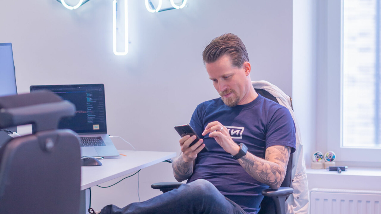 En man sitter vid ett skrivbord, han har benen i kors och tittar på sin mobiltelefon.