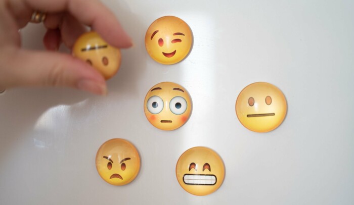 Emojis med olika uttryck på vit yta. Hand håller neutral emoji lite framför ytan.