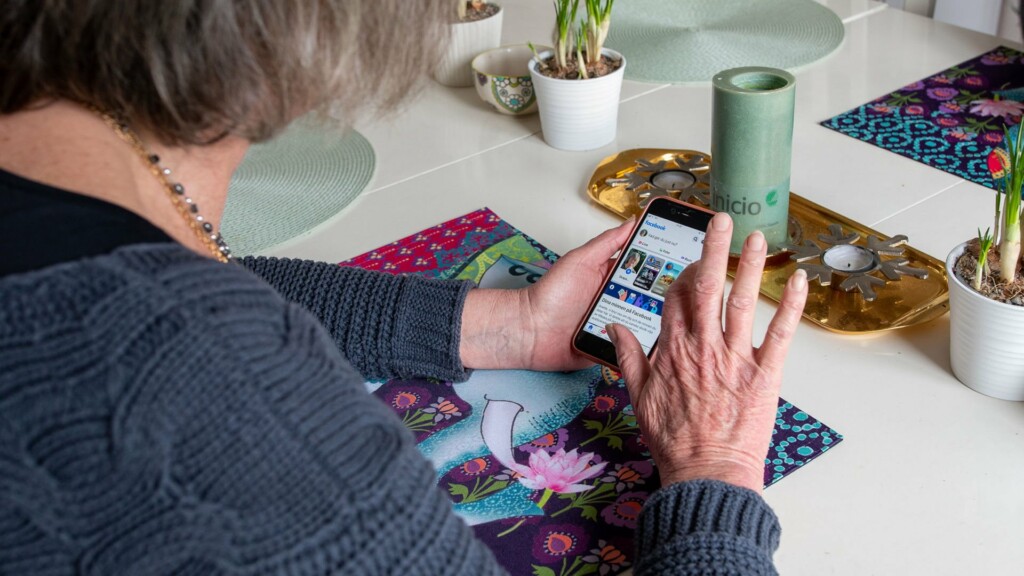 En äldre kvinna sitter vid köksbordet med mobilen i handen, på skärmen syns sociala medier.