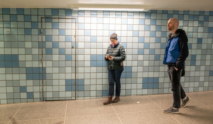 Äldre person står lutad mot en vägg och kollar sin mobil.