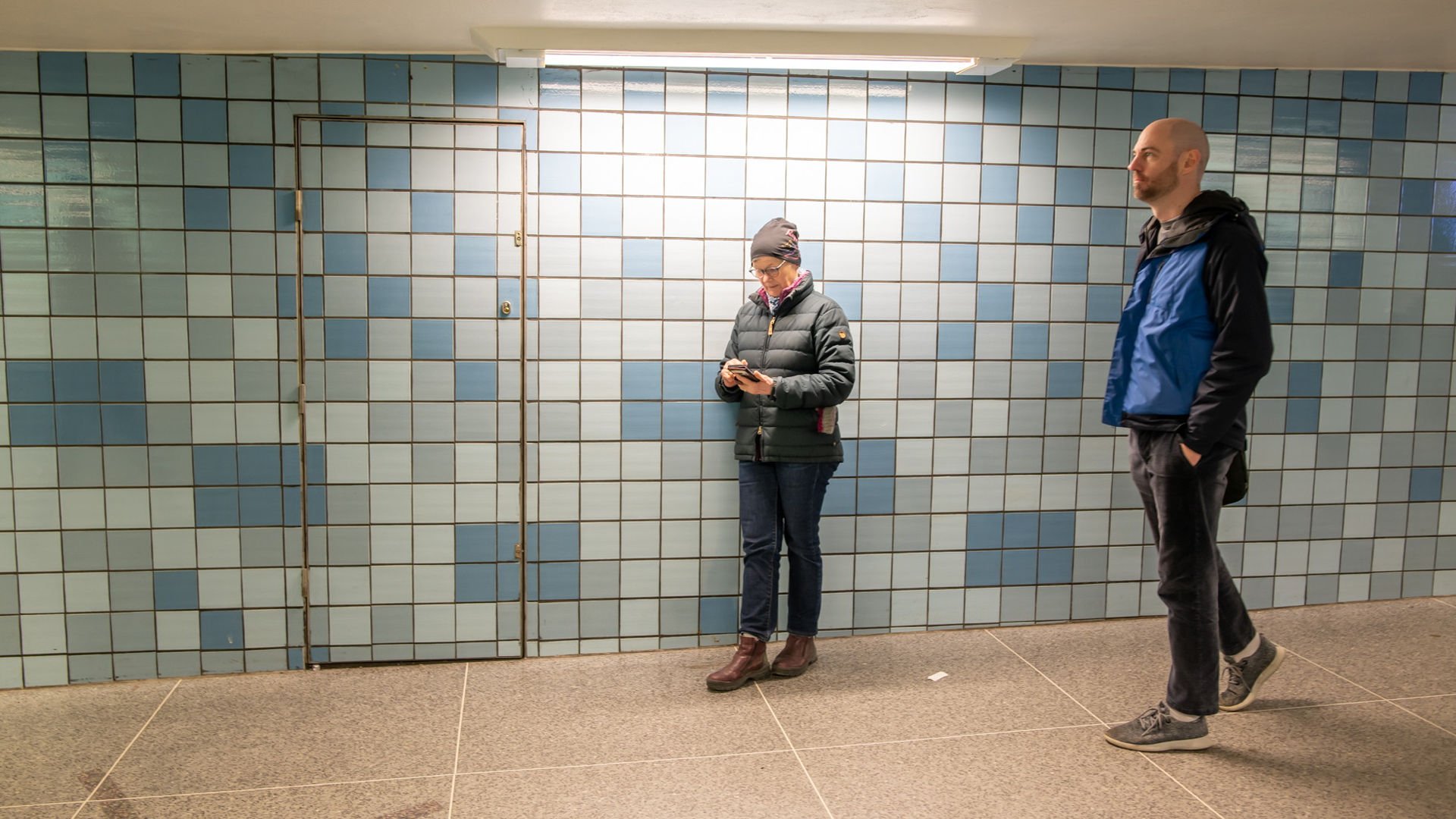 Äldre person står lutad mot en vägg och kollar sin mobil.