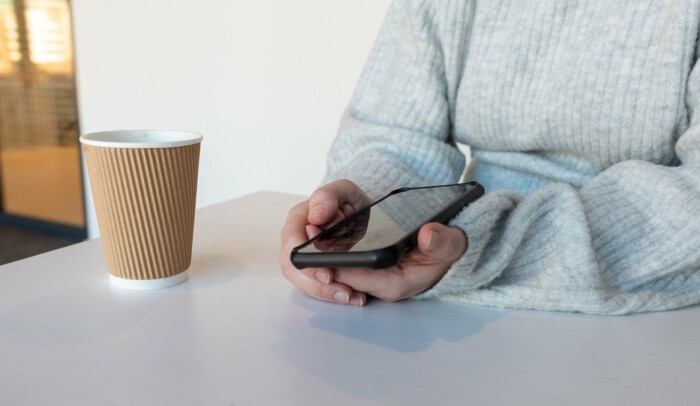 Kvinnliga händer som håller en mobiltelefon vid ett bord, bredvid står en pappersmugg.