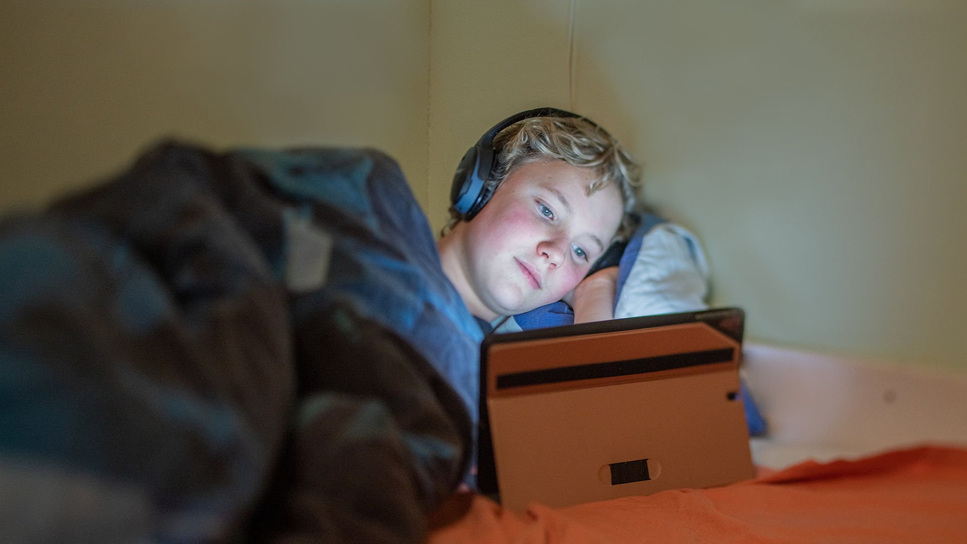 En pojke ligger i sängen med hörlurar och tittar på sin platta.