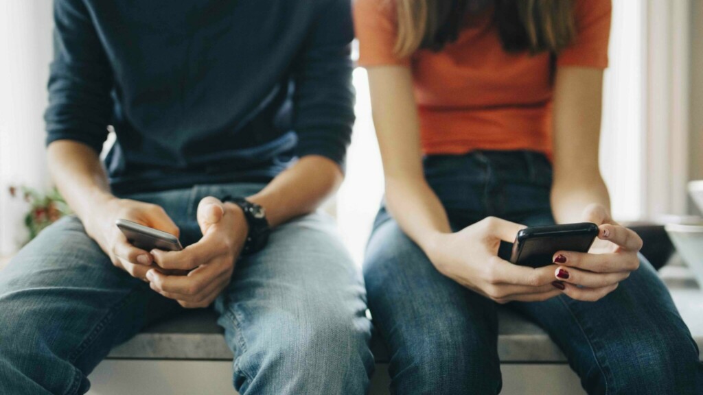 Två tonåringar sitter på köksbänk med varsin mobilelefon i knät. Prata nakenbilder och dickpics med din tonåring