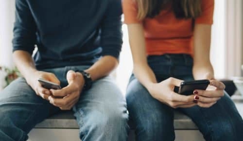 En tjej och en kille sitter på en bänk bredvid varandra och håller i varsin mobiltelefon