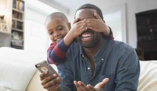 Pojke håller för ögonen på sin pappa som skrattar och håller mobiltelefon i handen.