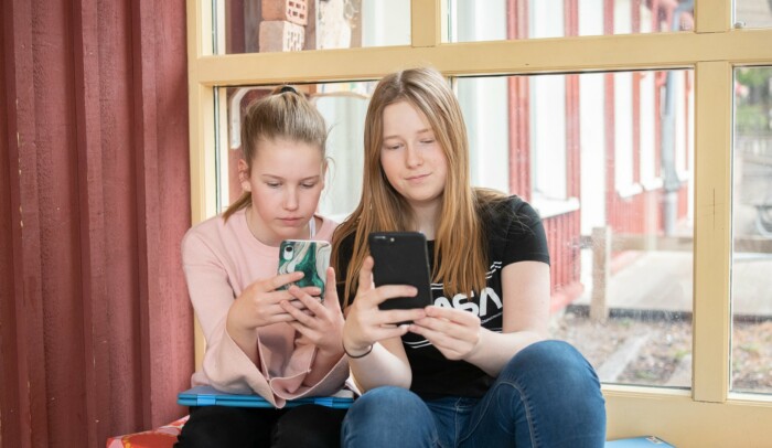 Två unga tjejer sitter i ett fönster med varsin mobil i handen.
