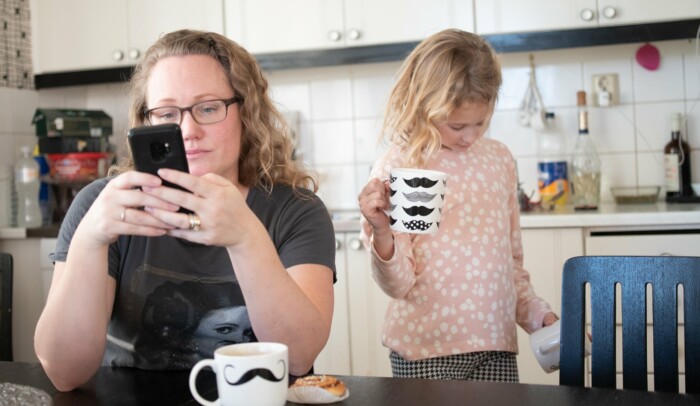 En kvinna sitter vid köksbordet och tittar på sin mobil, ett barn står bakom henne.