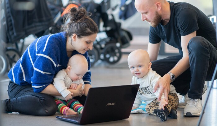 En kvinna och en man som håller om varsitt barn samtalar och använder en dator. Ena barnet tittar upp mot kameran.