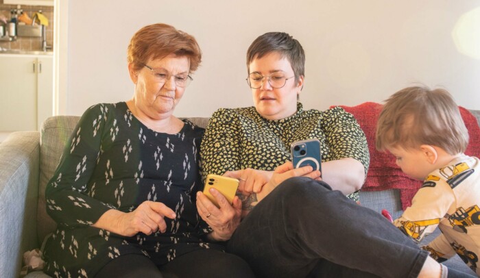 Två kvinnor sitter i soffa med varisn mobil i handen, vid sidan står ett barn och leker.