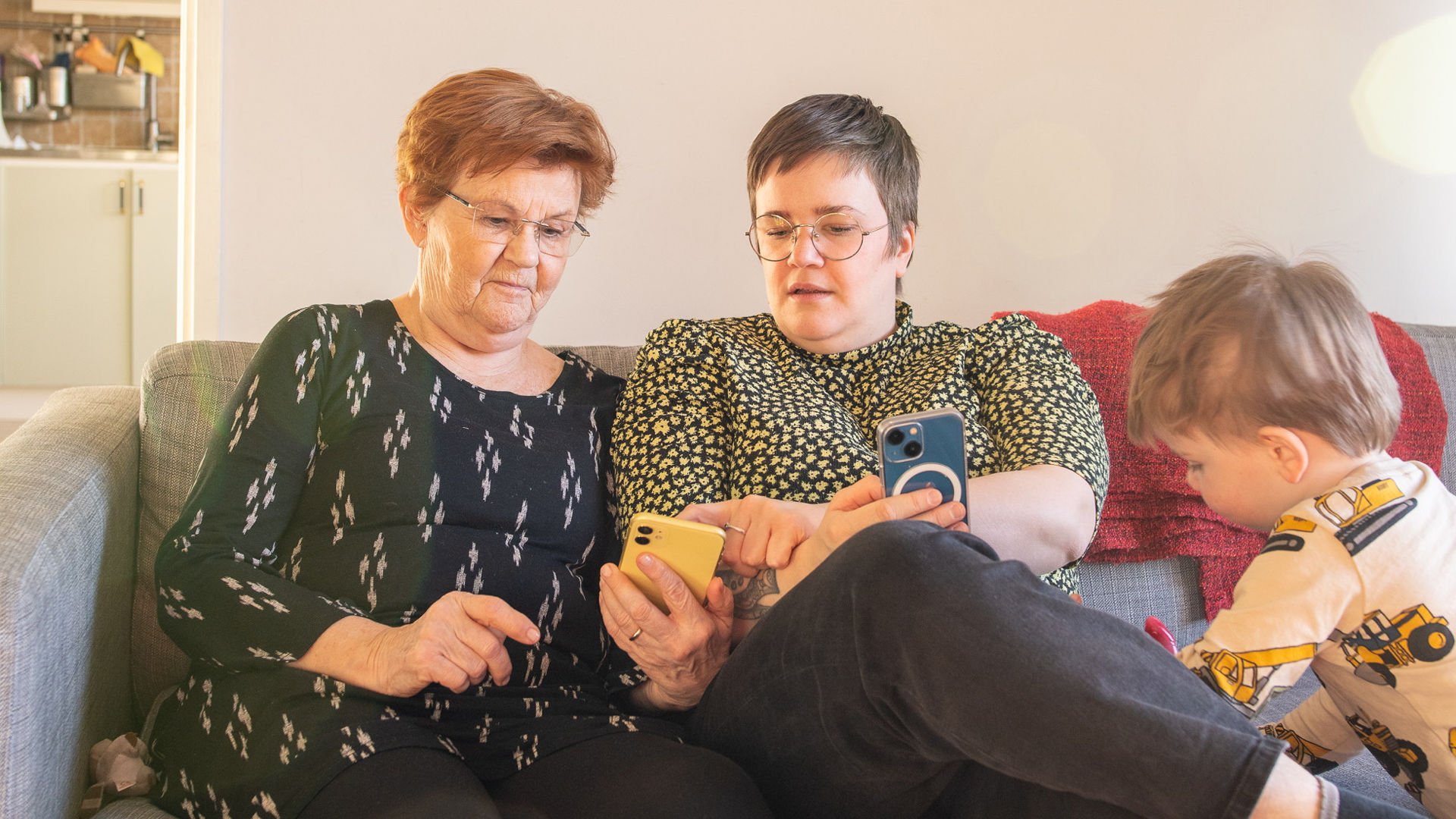 Två kvinnor sitter i soffa med varisn mobil i handen, vid sidan står ett barn och leker.