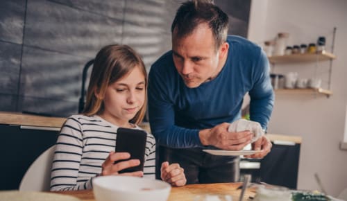 Flicka sitter vid köksbordet och visar sin pappa något i mobilen