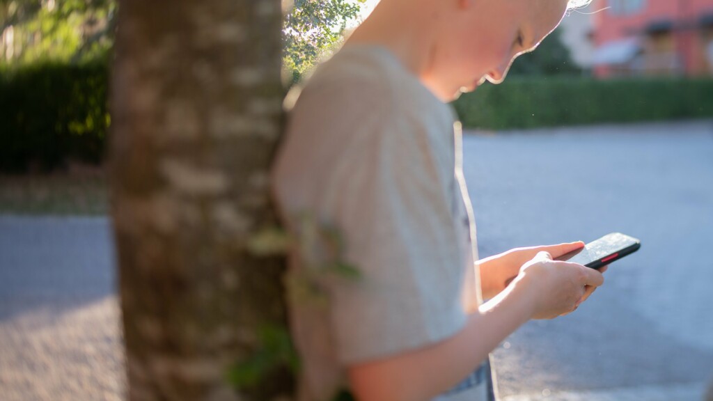 Barn som står lutat mot ett träd med mobilen i handen.