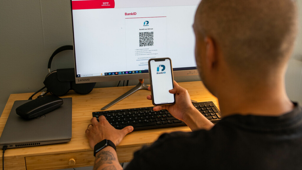En man sitter framför datorn med sin mobil i handen, på mobilen syns bank-id-appen.