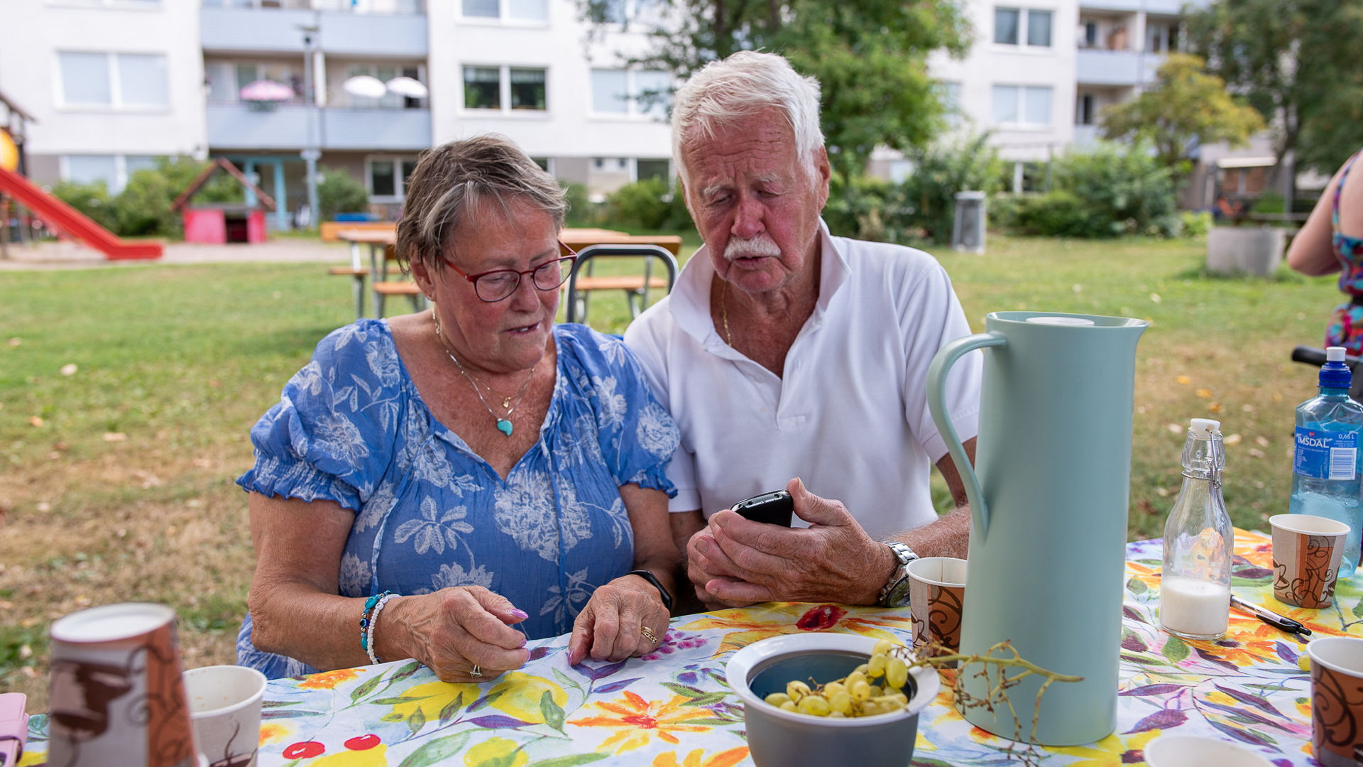 Äldre man och kvinna sitter vid ett bord utomhus, de tittar tillsammans på en mobil.