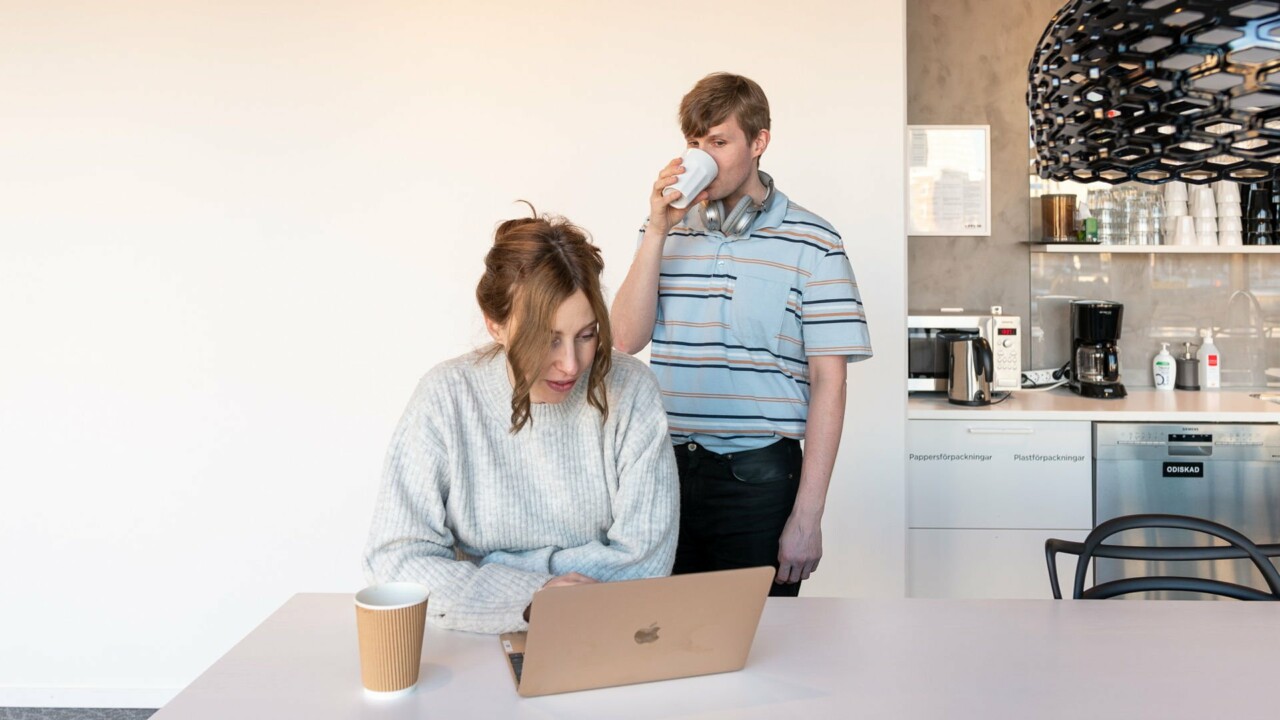 En kvinna sitter vid en laptop, en man står bakom henne och sneglar på skärmen