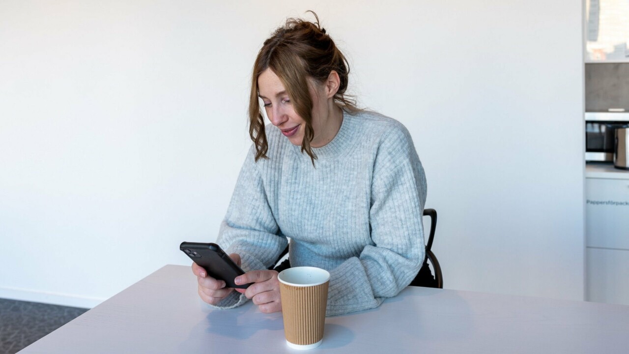 en kvinna sitter med sin mobil i handen och en kaffemugg framför sig
