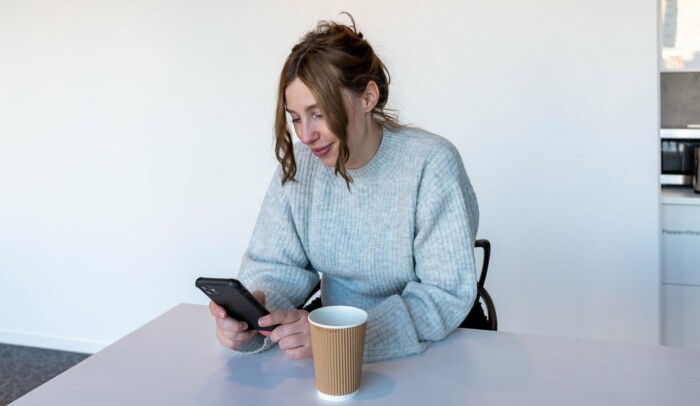 en kvinna sitter med sin mobil i handen och en kaffemugg framför sig