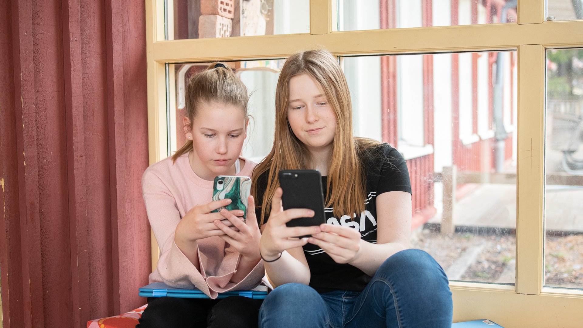 Två barn sitter och tittar på sina mobiler vid ett fönster.