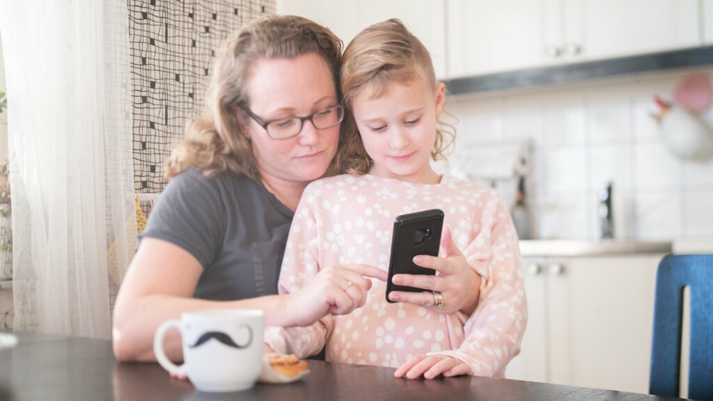 Kvinna vid köksbord använder mobiltelefon medan hon håller om ett barn.