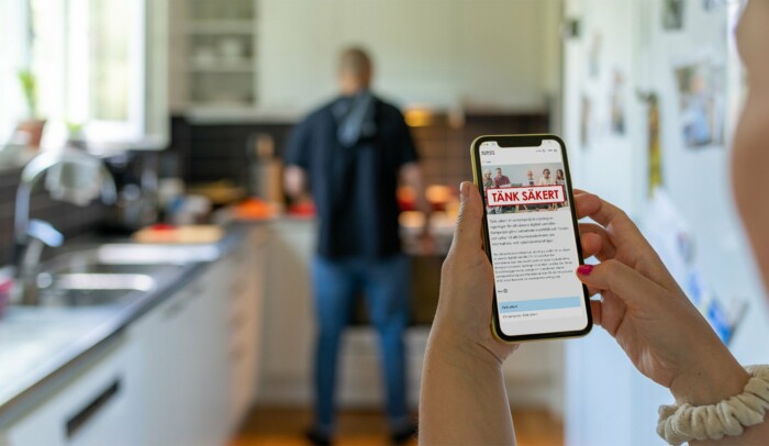 En man står i ett kök, i förgrunden håller någon i en mobil med Tänk säkert-kampanjen på skärmen.
