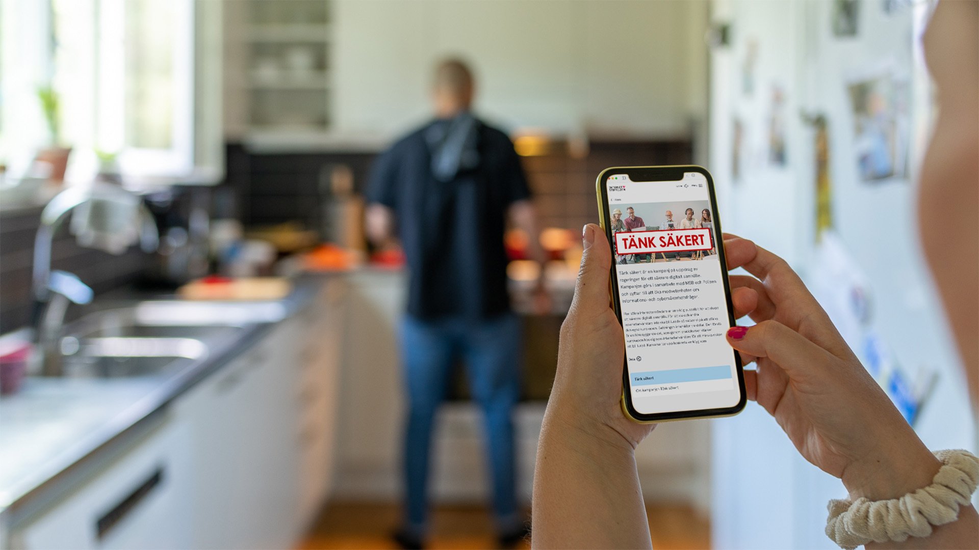 En man står i ett kök, i förgrunden håller någon i en mobil med Tänk säkert-kampanjen på skärmen.