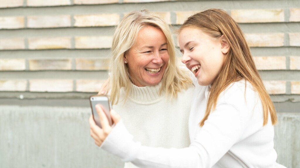 Kvinna och flicka, båda med vita tröjor, tar en bild tillsammans med en mobilkamera och skrattar.