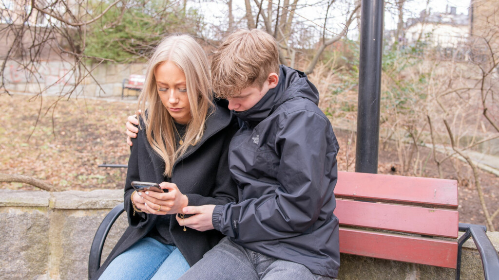 En ung kille håller om en ung tjej som ser ledsen ut, de sitter på en parkbänk och tittar på en mobil som tjejen håller i sina händer.