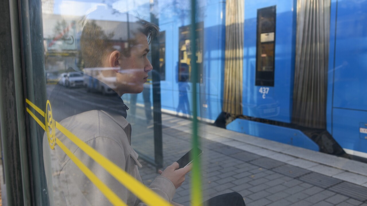 En ung kille sitter i en kur vid tvärbanan och tittar på sin mobil.
