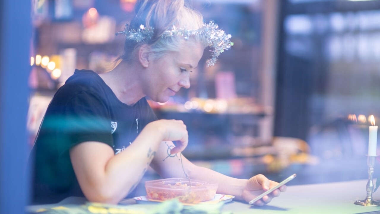 En kvinna sitter på ett cafe och scrollar i sin mobil, hon har glitter i håret och ser glad ut.