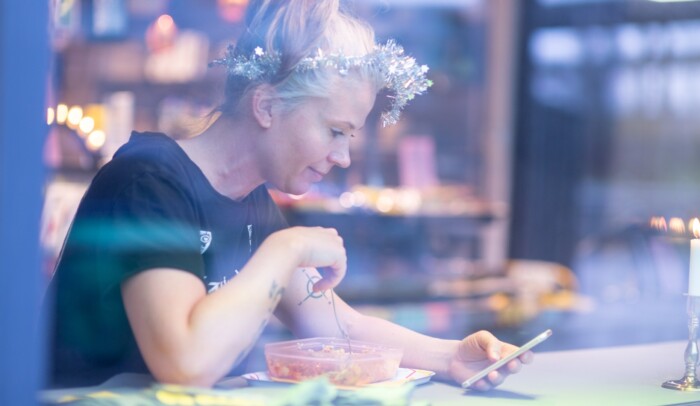 En kvinna sitter på ett cafe och scrollar i sin mobil, hon har glitter i håret och ser glad ut.