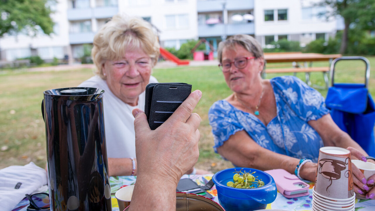 Två äldre kvinnor tittar på en mobilskärm som någon annan håller upp framför dem, de är utomhus.