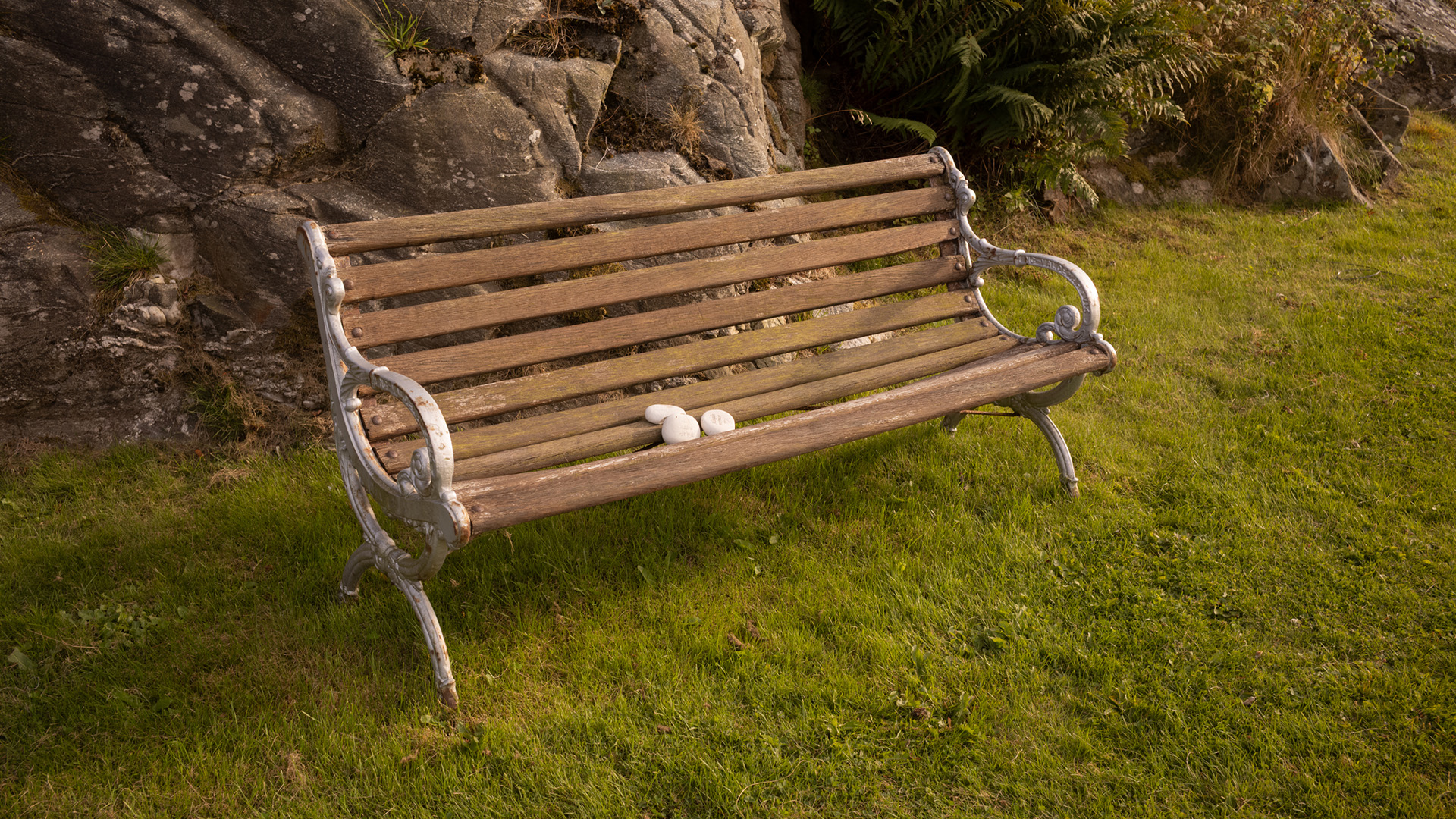 En parkbänk står på en gräsmatta på en kyrkogård. På bänken ligger tre små vita stenar.