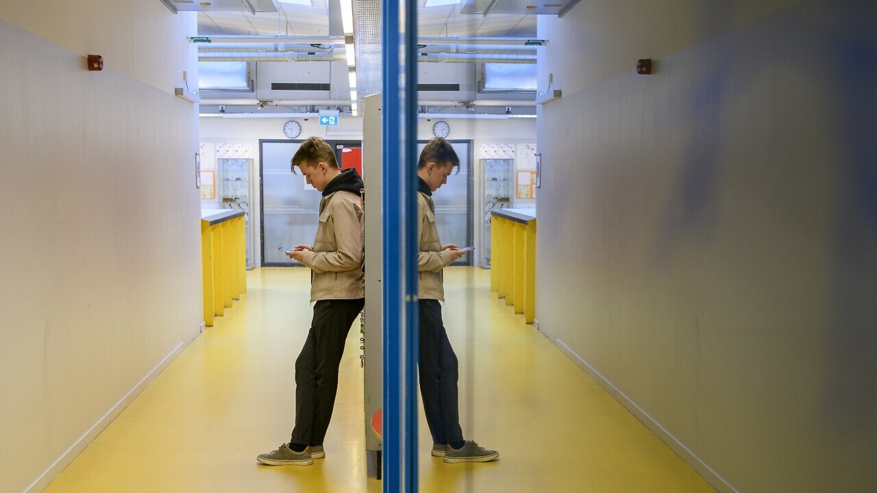 En ung kille som står i en korridor i skolan och tittar på mobilen.